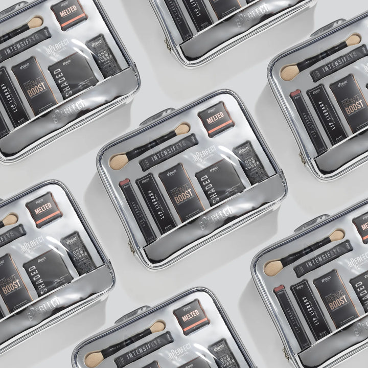 Multiple Makeup products including brushes, palettes, and a spray bottle in a clear cosmetic bag on a light gray background.