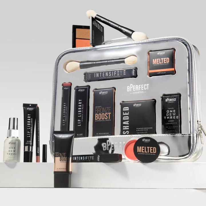 Makeup products including brushes, palettes, and a spray bottle in a clear cosmetic bag on a light gray background.