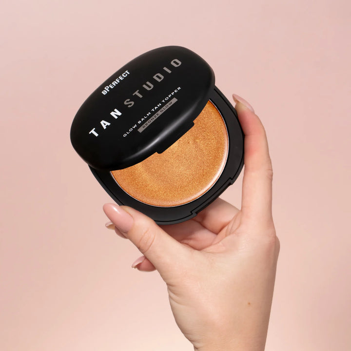 Hand holding a compact tan topper with 'TAN STUDIO' branding on a pink background