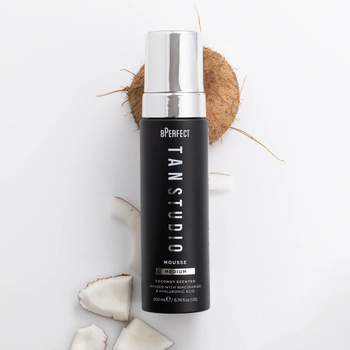 Black bottle of BPerfect Tan Studio Mousse with silver cap and coconuts on a white background