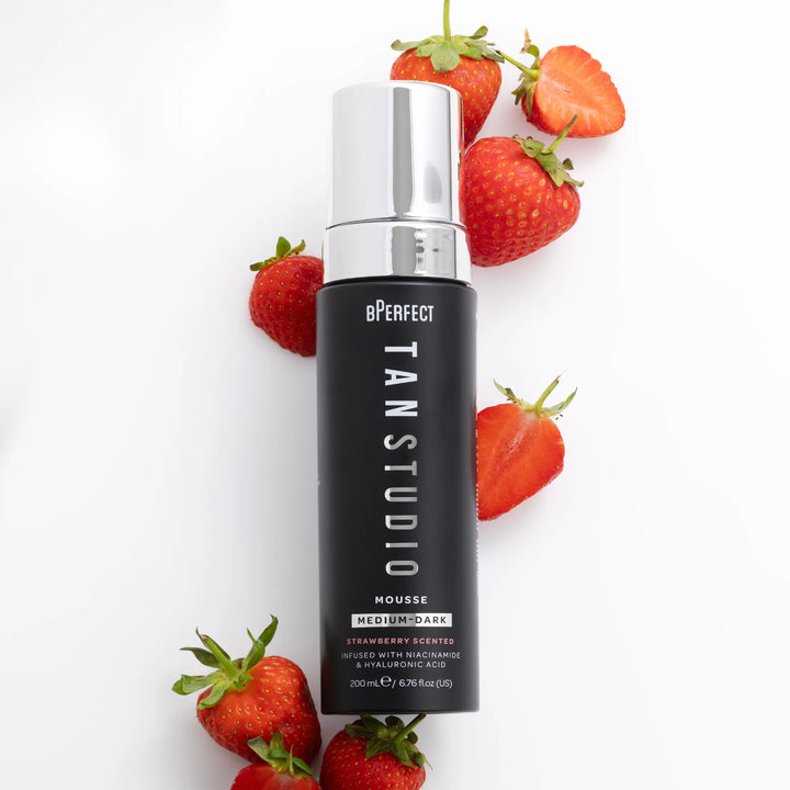 Black bottle of BPerfect Tan Studio Mousse with silver cap and strawberries on a white background